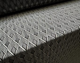 May include: Close-up of a textured fabric with a repeating geometric pattern. The design features diamond shapes with intricate lines and small square details, in shades of gray and white. The fabric appears to be folded over.