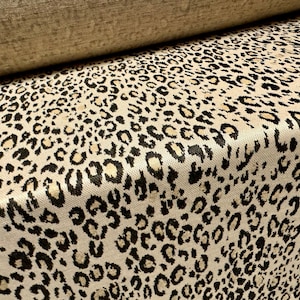 Op de afbeelding: Close-up van een beige stof met een zwart en gouden luipaardprint. De stof heeft een textuur.