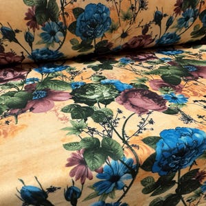 May include: Floral fabric with a repeating pattern of large blue, purple, and green flowers on a light orange background. The fabric has a soft, textured appearance, with detailed botanical elements.