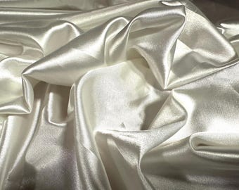 May include: Close-up of ivory-colored satin fabric with soft folds and a lustrous sheen. The fabric's surface reflects light, creating highlights and shadows. This textile is suitable for apparel, home decor, or craft projects.