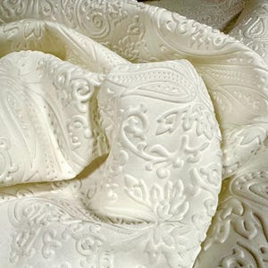 Peut inclure: Gros plan sur un tissu blanc avec un motif cachemire en relief. Le tissu est plié et drapé, mettant en valeur le motif en relief complexe. La texture et les détails suggèrent un textile luxueux, peut-être pour la décoration intérieure ou la mode.