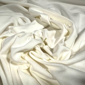 Op de afbeelding: Close-up van een witte stof met een zachte, gladde textuur. De stof is gevouwen en gedrapeerd, waardoor een gevoel van beweging en stroming ontstaat.