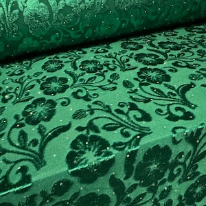 May include: Close-up of emerald green fabric with a floral design. The fabric has a velvet texture and a repeating pattern of flowers and leaves. Tiny, sparkling embellishments are scattered across the surface, adding a subtle shimmer.