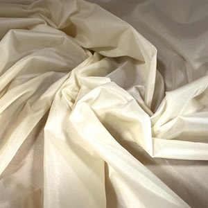 Puede incluir: Primer plano de una tela de color crema con una textura suave y sedosa. La tela está drapeada y doblada, creando líneas suaves y fluidas.