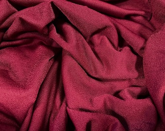 Może przedstawiać: Zbliżenie na głęboki bordowy materiał o fakturowanej powierzchni. Materiał jest złożony i udrapowany, tworząc miękkie, płynne linie.