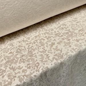 Op de afbeelding: Close-up van een witte stof met een subtiel beige patroon. De stof heeft een textuur, een licht ruw oppervlak.