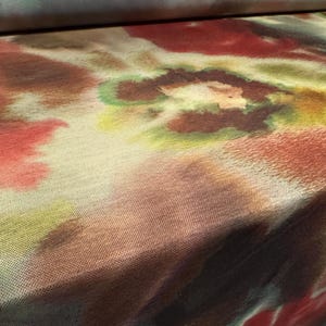 Könnte beinhalten: Nahaufnahme eines Stoffes mit einem aquarellartigen Design. Der Stoff weist eine Mischung aus Farben auf, darunter Rot, Braun, Grün und Gelb, wodurch ein weiches, strukturiertes Aussehen entsteht. Das Muster ähnelt abstrakten floralen oder organischen Formen.