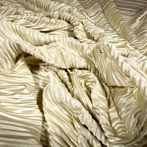 May include: Close-up of a cream-coloured fabric with a pleated texture. The fabric is draped and folded, creating a sense of depth and movement. The surface has a subtle sheen, reflecting light and highlighting the ridges.