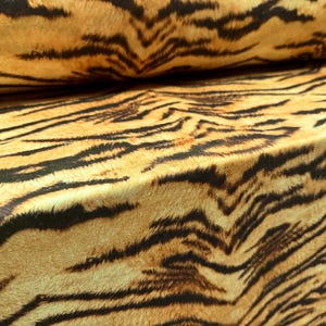 May include: Close-up of a fabric with a tiger stripe pattern. The fabric has a tan background with black stripes. The fabric is folded over, showing the texture and pattern of the material.