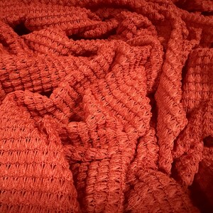 May include: Close-up of a textured, coral-coloured fabric. The material is intricately woven with a repeating pattern, creating a visually interesting surface. The fabric is draped, showing folds and shadows.