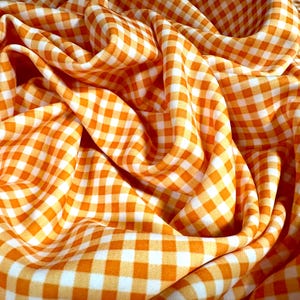 Könnte beinhalten: Nahaufnahme eines orange-weiß karierten Stoffes. Der Stoff ist drapiert und gefaltet, wodurch weiche Kurven und Schatten entstehen. Das Muster ist ein klassisches Karomuster mit orangefarbenen und weißen Quadraten. Der Stoff scheint leicht und für verschiedene Nähprojekte geeignet zu sein.