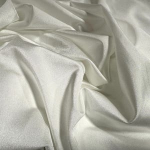 Pode incluir: Close-up de tecido branco com dobras macias e textura sutil. O tecido parece ser um material leve e fluido, possivelmente seda ou um têxtil semelhante. As dobras criam sombras e destaques, adicionando profundidade à imagem.