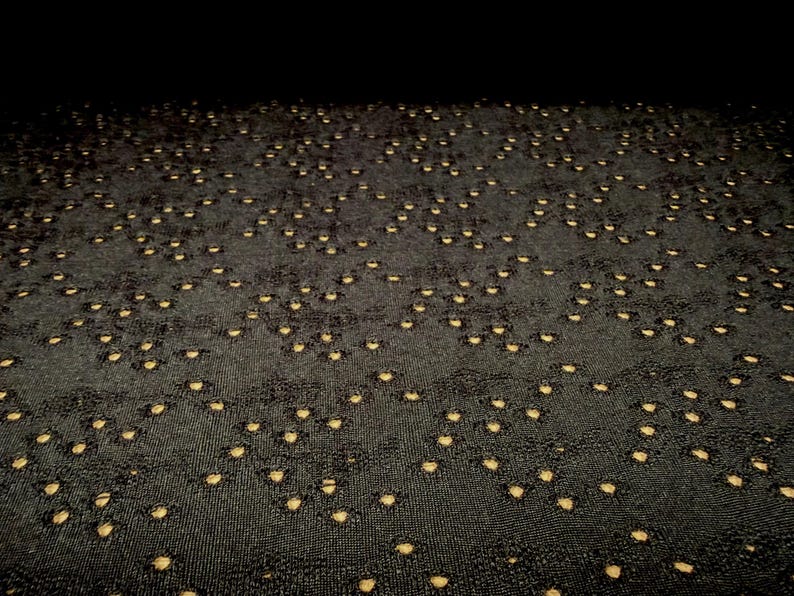 May include: A black fabric featuring a repeating pattern of small, circular cutouts that reveal a light beige or tan layer underneath. The material has a textured, woven appearance with a consistent grid of perforations across the entire surface.