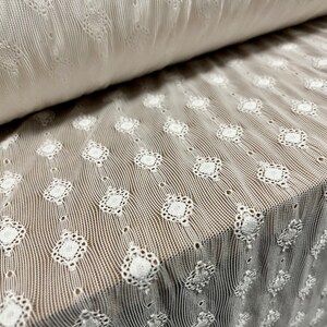 May include: White cotton fabric with a delicate, embroidered pattern of small, repeating diamond shapes. The fabric has a slightly textured, crinkled look.