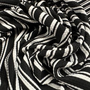 May include: Black and white striped fabric with a textured, crinkled look. The fabric is folded and draped, showing the pattern and texture.