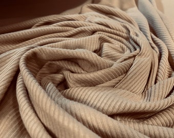 May include: Close-up of a soft, beige ribbed fabric. The fabric is folded and twisted, creating a swirling pattern.