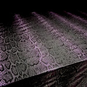 Puede incluir: Una tela con un patrón de piel de serpiente texturizado y en relieve en tonos negro y morado oscuro. El material muestra un diseño repetitivo de escamas de reptil con un acabado mate, creando una superficie táctil distintiva y definida.
