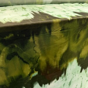 May include: Close-up of fabric with a watercolor-like design. The fabric features a blend of green, brown, and white hues, creating a textured, abstract pattern. The material appears to be folded, showcasing the design's flow and depth. Ideal for apparel or home decor.
