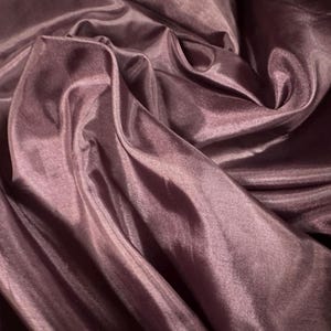 May include: A close-up shot of a mauve-coloured fabric with a shimmering finish. The material is draped, showing soft folds and highlights. The fabric's texture suggests it could be used for clothing or decorative items, with a flowing, lightweight quality.