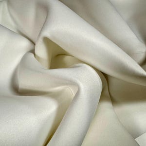 Könnte beinhalten: Nahaufnahme eines elfenbeinfarbenen Stoffes mit weichen Falten und subtilen Schatten. Der Stoff scheint glatt zu sein, möglicherweise Satin oder Seide, mit leichtem Glanz. Die Falten erzeugen ein Gefühl von Tiefe und Textur, was auf ein luxuriöses Textil hindeutet.