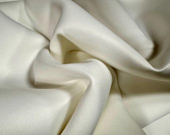 Puede incluir: Primer plano de una tela de color marfil con pliegues suaves y sombras sutiles. La tela parece lisa, posiblemente satén o seda, con un ligero brillo. Los pliegues crean una sensación de profundidad y textura, sugiriendo un textil lujoso.