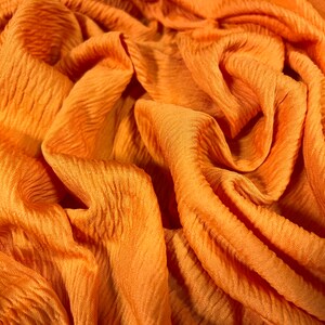 May include: Close-up of a textured orange fabric with a crinkled, wrinkled appearance. The fabric has a soft, flowing drape.