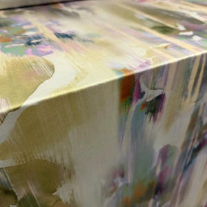 Op de afbeelding: Close-up van een stof met een abstract aquarelontwerp. De stof heeft een mix van kleuren, waaronder geel, groen, paars en roze, waardoor een zachte, artistieke uitstraling ontstaat. De randen van de stof zijn zichtbaar en tonen de dikte en textuur.