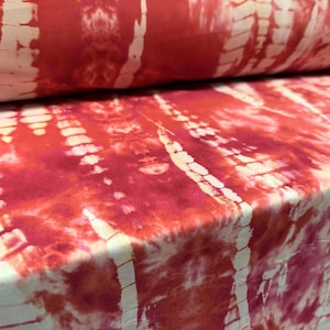 Könnte beinhalten: Nahaufnahme eines Stoffes mit einem Batikmuster in Rot- und Weißtönen. Der Stoff ist gefaltet und zeigt die Textur und das Design. Das Muster weist vertikale Streifen und einen Aquarelleffekt auf.