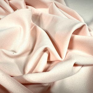 Könnte beinhalten: Nahaufnahme eines weichen, rosafarbenen Stoffes mit strukturierter Oberfläche. Der Stoff ist drapiert und gefaltet, wodurch Schatten und Lichtreflexe entstehen, die seine Textur und seinen Fluss betonen. Das Gesamtbild ist glatt und zart.