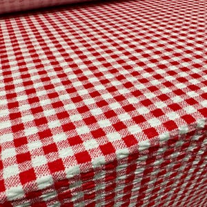Könnte beinhalten: Nahaufnahme eines rot-weiß karierten Stoffes. Der Stoff ist gefaltet und zeigt einen Stapel Material. Das Muster ist ein klassisches Karomuster mit gleichmäßig verteilten Quadraten. Der Stoff scheint ein mittelschweres Material zu sein.