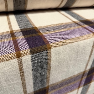 May include: Close-up of a fabric with a plaid pattern. The fabric features a cream-coloured base with intersecting lines in shades of purple, brown, and grey. The fabric is folded over, revealing the pattern's repeat.