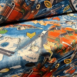 May include: Close-up of a fabric with a denim-like background, featuring various decorative elements. The design includes zippers, safety pins, watches, and patches. The fabric also has a plaid pattern in shades of orange and red.