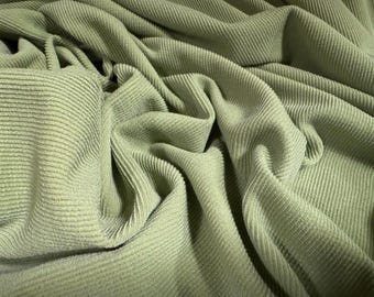 May include: Close-up of sage green corduroy fabric. The fabric is draped and folded, showcasing the ribbed texture. The color is a muted, earthy green, and the fabric appears soft and pliable.