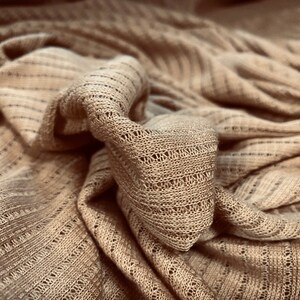 Könnte beinhalten: Nahaufnahme eines beigefarbenen Strickstoffes mit einem strukturierten, offenen Webmuster. Der Stoff ist gefaltet und drapiert und erzeugt einen weichen und gemütlichen Look.