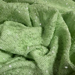 Puede incluir: Primer plano de una tela verde claro cubierta de pequeñas lentejuelas redondas y brillantes. La tela está drapeada y doblada, creando sombras y texturas. Las lentejuelas capturan la luz, agregando un brillo sutil. El efecto general es suave y lujoso.