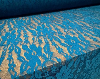 Pode incluir: Close-up de tecido de renda azul-petróleo com um padrão floral. A renda é sobreposta a um tecido bege, criando um contraste. O tecido está enrolado ao fundo.