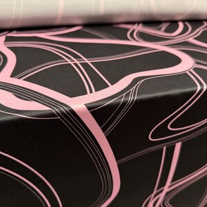 May include: Close-up of black wrapping paper with a pink abstract design. The design features flowing, curved lines and swirls. A roll of paper with a similar design in a lighter color is visible in the background.