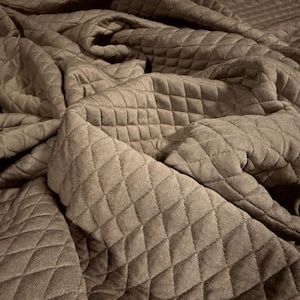 Puede incluir: Primer plano de una manta acolchada marrón. La manta tiene un patrón de diamantes y está doblada y drapeada, creando sombras y texturas. La tela parece suave y acogedora, adecuada para ropa de cama o decoración del hogar.