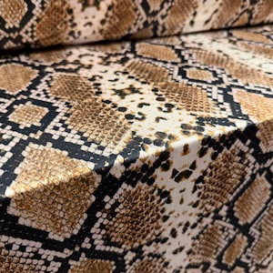 May include: Close-up of a fabric with a snakeskin pattern. The material displays a repeating design of brown, tan, and black scales, creating a realistic reptile skin texture. The fabric is folded, showing the pattern's continuity.
