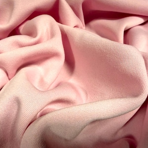 Puede incluir: Primer plano de una tela rosa claro, suave y con textura, con pliegues visibles. El material parece ser de punto o jersey, con un ligero brillo. La imagen destaca la superficie lisa y las suaves curvas de la tela.