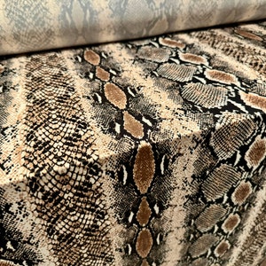 May include: A close-up of a fabric with a snakeskin pattern. The fabric has a repeating design of brown, tan, and black scales. A roll of grey fabric is visible in the background, suggesting a textile product.