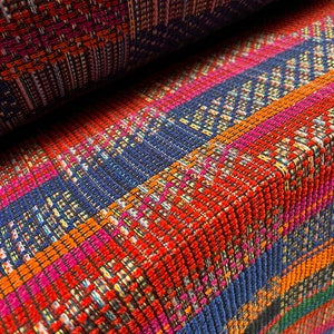 Könnte beinhalten: Nahaufnahme eines gewebten Stoffes mit einem lebendigen Streifenmuster. Das Design zeigt Bänder in Rot, Orange, Blau, Pink und Grün. Die Textur des Stoffes ist sichtbar, mit leichtem Glanz. Der Stoff ist gefaltet und zeigt die Unterseite.