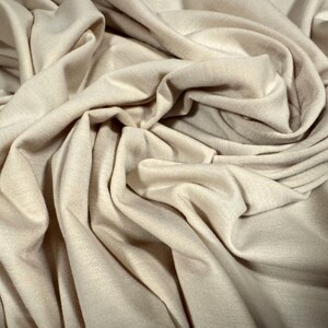 Pode incluir: Um close-up de um tecido bege com uma textura macia e lisa. O tecido está drapeado e dobrado, criando um visual luxuoso e elegante.