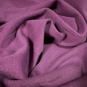 Pode incluir: Um close-up de um tecido de veludo cotelê roxo escuro com uma superfície texturizada e canelada. O tecido está dobrado e drapeado, criando dobras e curvas suaves.