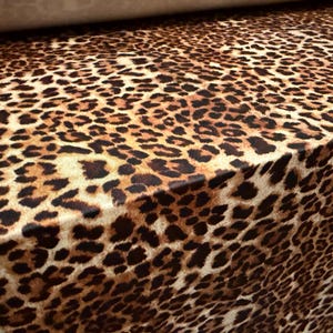 May include: Close-up of a fabric with a leopard print pattern. The fabric features a tan background with dark brown and black spots. The fabric is folded over, showing the pattern's repeating design.