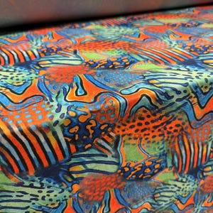Könnte beinhalten: Nahaufnahme eines Stoffes mit einem lebendigen, abstrakten Muster. Das Design zeigt eine Mischung aus orangefarbenen, blauen und grünen Formen mit gepunkteten und gestreiften Details. Der Stoff scheint weich und fließend zu sein, mit leichtem Glanz.