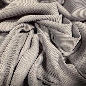 May include: Close-up of grey fabric with a ribbed texture. The fabric is draped and folded, creating shadows and depth. The material appears soft and has a subtle sheen, suggesting it could be used for clothing or home decor.