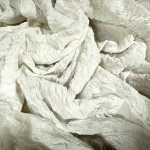Puede incluir: Tela blanca texturizada con un aspecto suave y arrugado. La tela está drapeada y doblada, creando una sensación de movimiento y textura.
