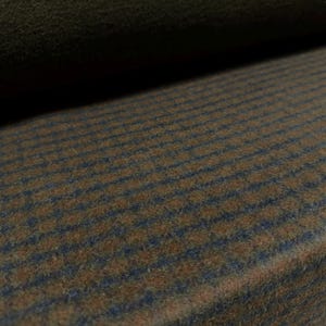Op de afbeelding: Een rol stof met een gestructureerd oppervlak en een herhalend patroon van dunne, donkerblauwe horizontale strepen op een warme, middenbruine achtergrond. Het materiaal heeft een zacht, licht pluizig uiterlijk met een dichte weving.