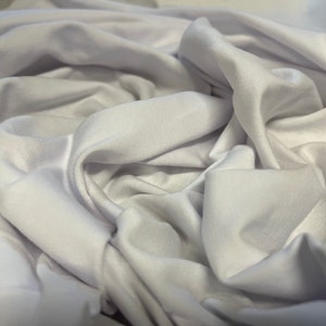 May include: White fabric with a smooth, silky texture. The fabric is folded and draped, creating soft, flowing folds.